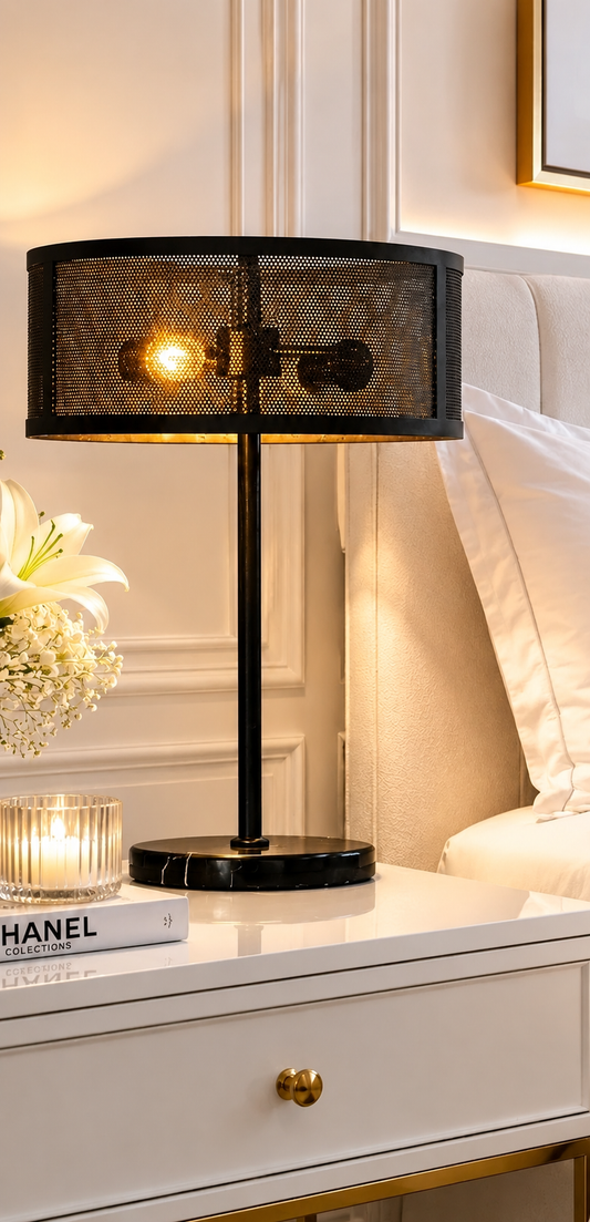 A black metal mesh table lamp with a warm glowing bulb, styled on a white bedside table next to a candle and a book.